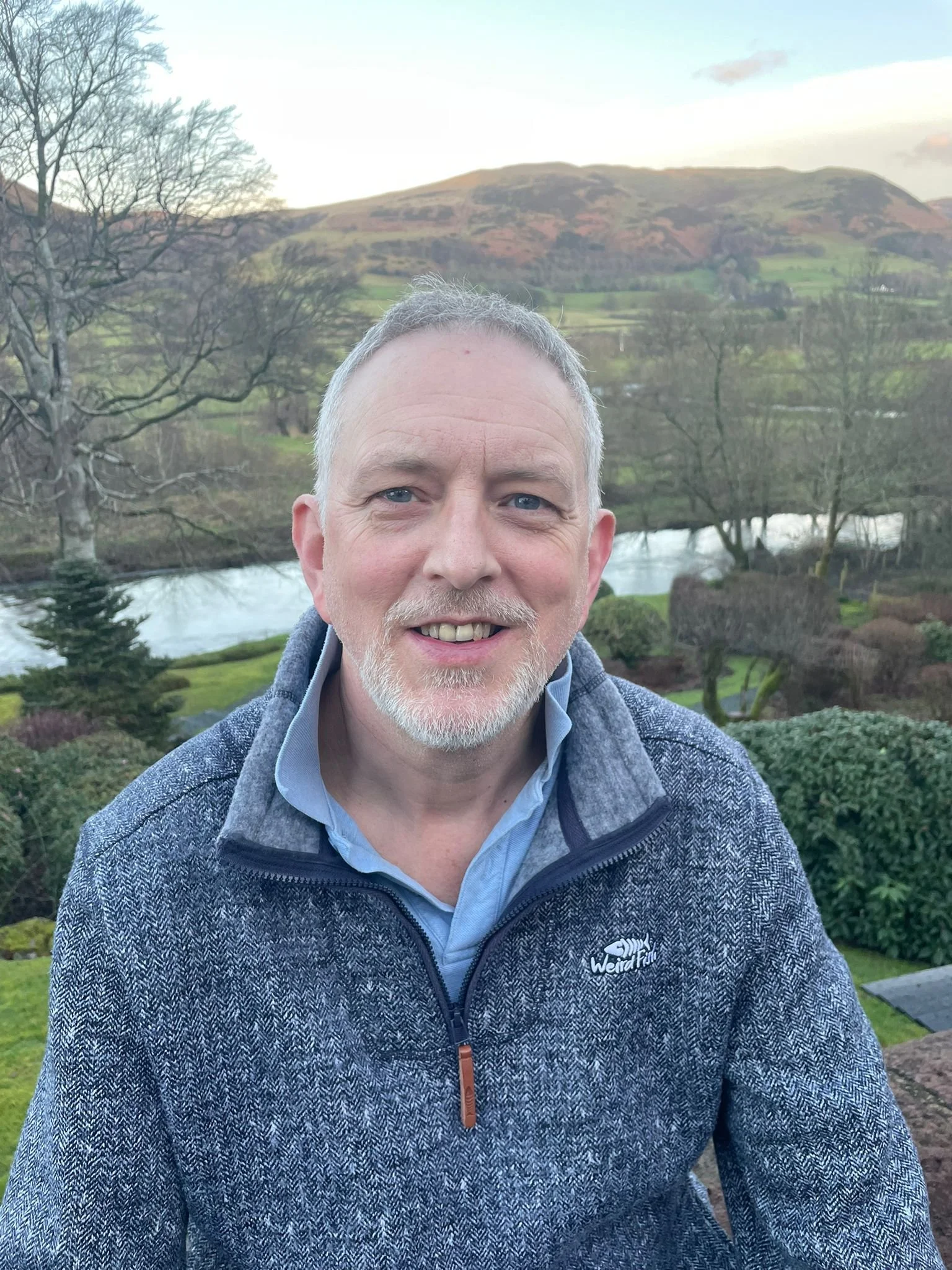 A smiling older man with gray hair and a beard, dressed in a blue shirt and a gray zip-up jacket, sits outdoors with a scenic background of trees, a river, and rolling hills on a clear day.