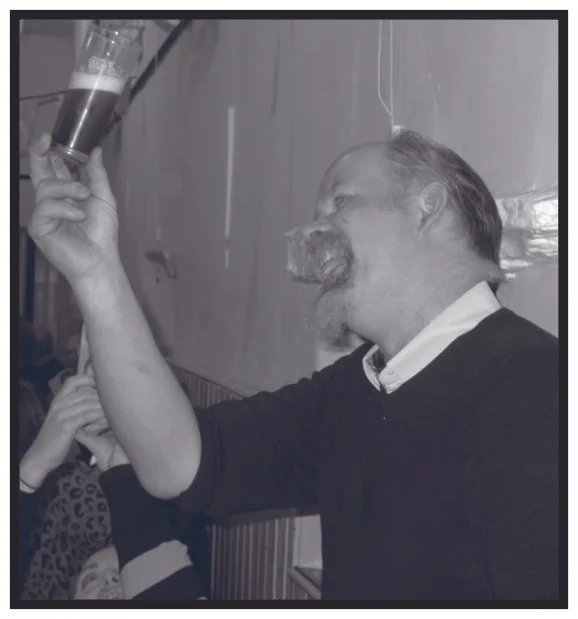 A man with a beard and mustache in a dark sweater holding a glass bottle, pouring or about to pour a drink, with another person partially visible in the background.