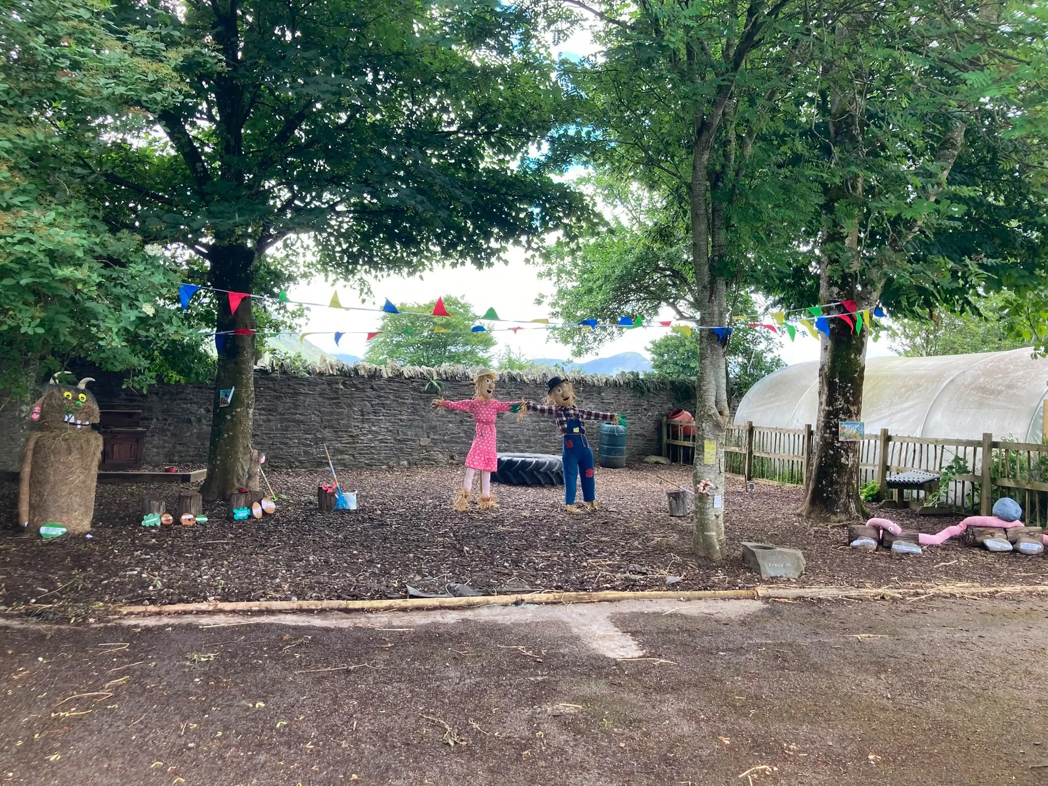 Outdoor scene with trees, bunting flags, scarecrow figures, and decorative items on the ground, possibly in a garden or park area.