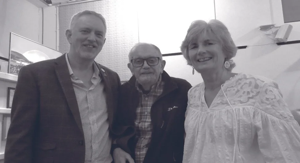 Three people standing together indoors, smiling for the camera. The person on the left is a young man in a suit jacket, the middle person is an elderly man with glasses, and the person on the right is a woman wearing a white lace blouse.