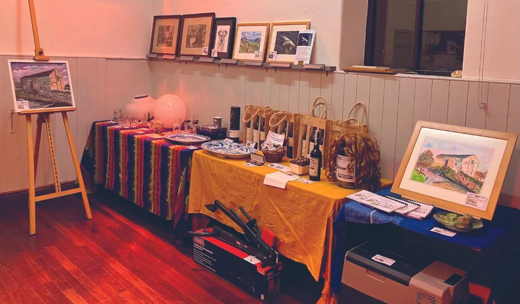 Art gallery display with framed landscape paintings on a wall shelf, a easel with a painting, and a table with various items including bottles, glasses, and artwork, covered with colorful cloths.