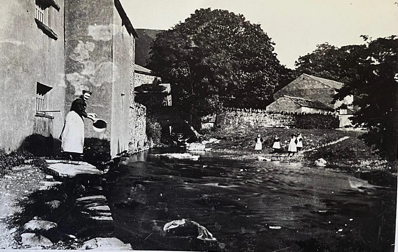A black-and-white photo showing a rural scene with a woman washing clothes by a water stream on the left, three girls walking along the stream on the right, and two buildings with trees in the background.