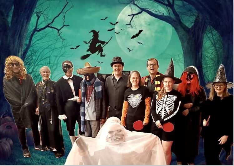 Group of people in costumes standing outdoors at night, with a full moon and spooky trees in the background, celebrating Halloween.