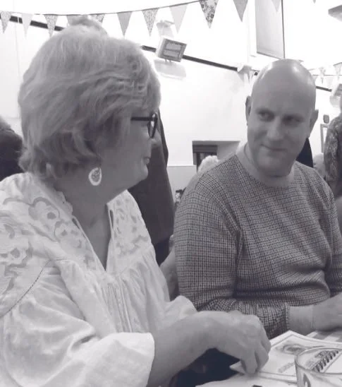 An elderly woman with glasses and earrings talking to a bald man with a checkered shirt at a gathering with bunting banners.