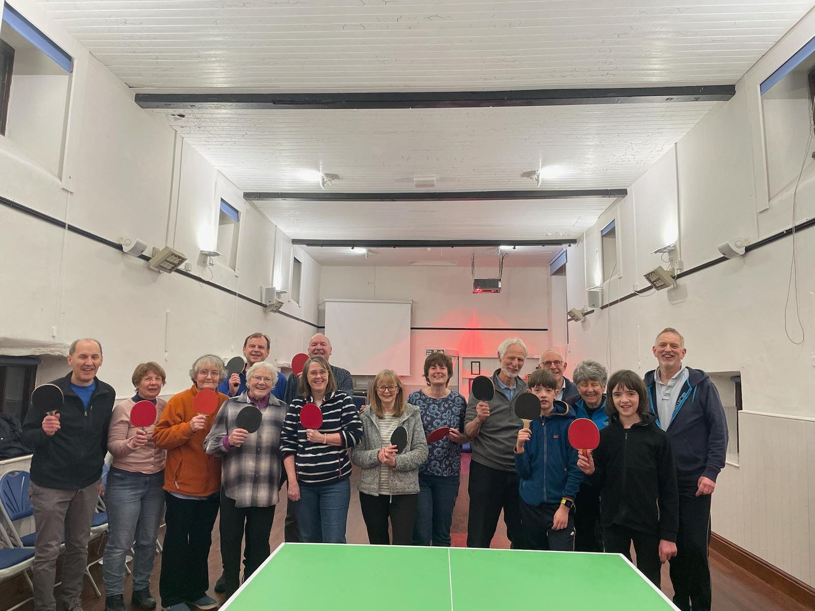 Group of people in a room with table tennis paddles, smiling for a photo, some children and adults, indoor setting with white walls and ceiling.