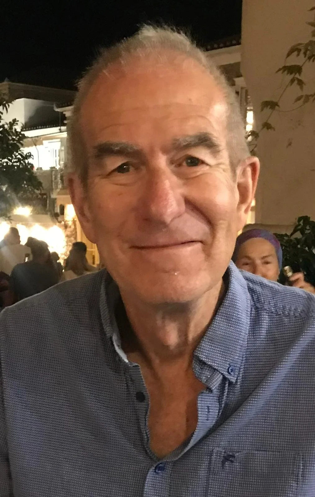 A middle-aged man with short, gray hair and a slight smile, wearing a blue button-up shirt, at an outdoor event at night.