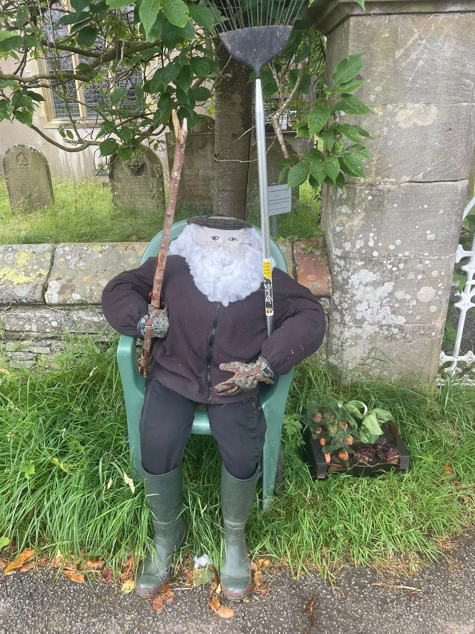 An outdoor scene featuring a scarecrow with a painted face, a white beard, a hat, gloves, and waterproof boots. The scarecrow is seated in a green plastic chair amid grass, plants, and flowers. There are trees, a stone wall, and old tombstones in the