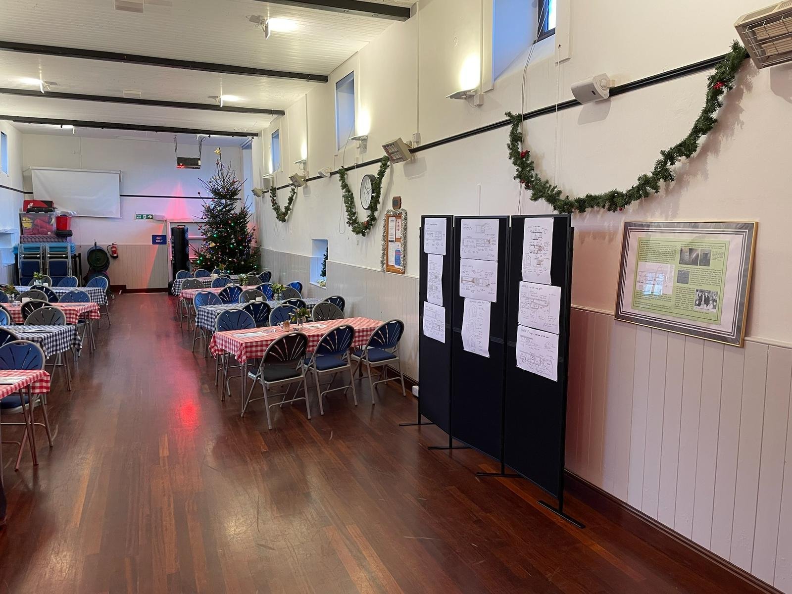 A decorated indoor space for a Christmas celebration featuring a Christmas tree with lights, tables with red and white checkered tablecloths, and holiday decorations on the wall including green garlands.