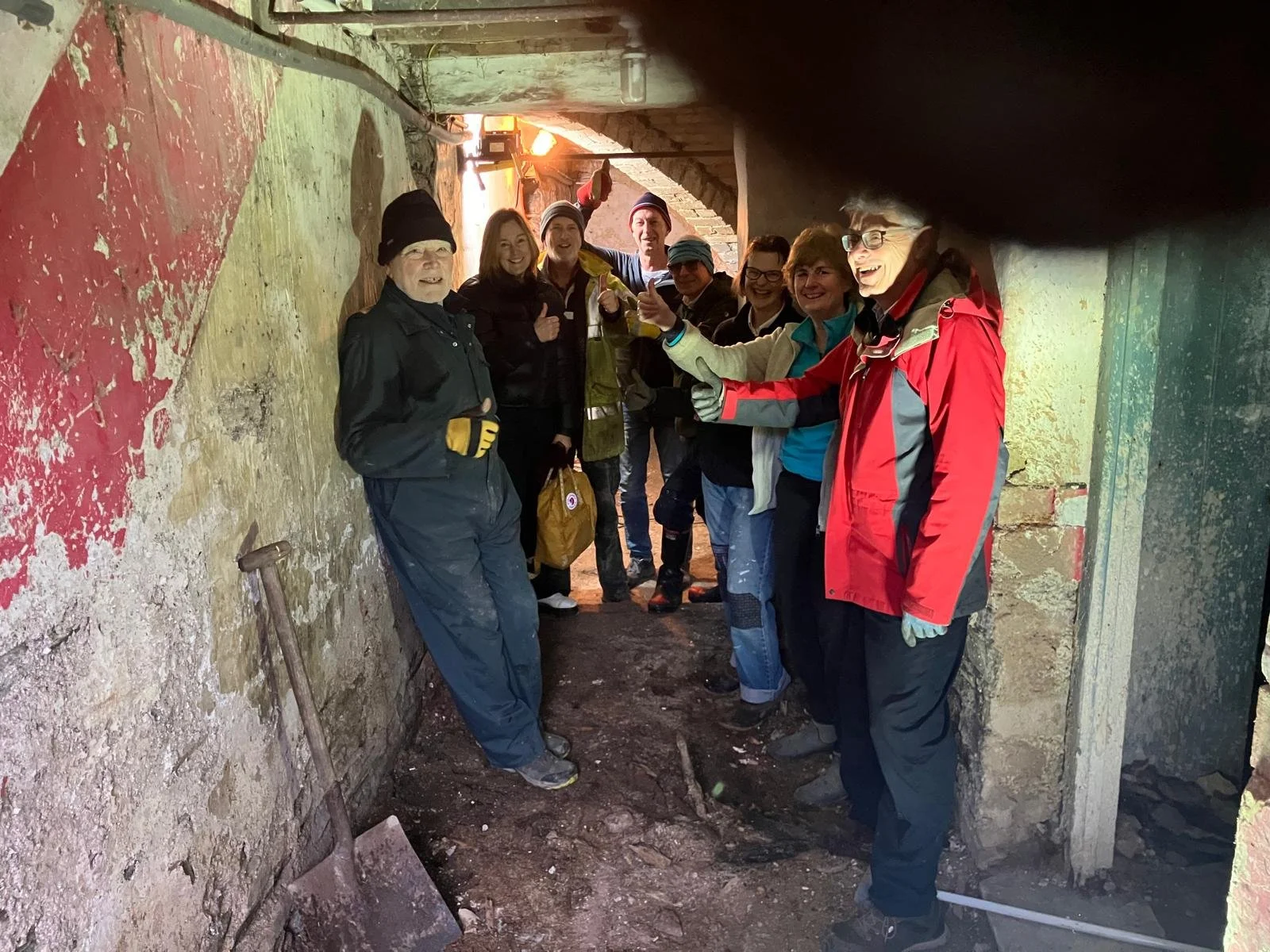 Group of people bundled in jackets and gloves inside a tunnel, smiling, giving thumbs up, construction tools like a shovel leaning against a wall, some people wearing hats.