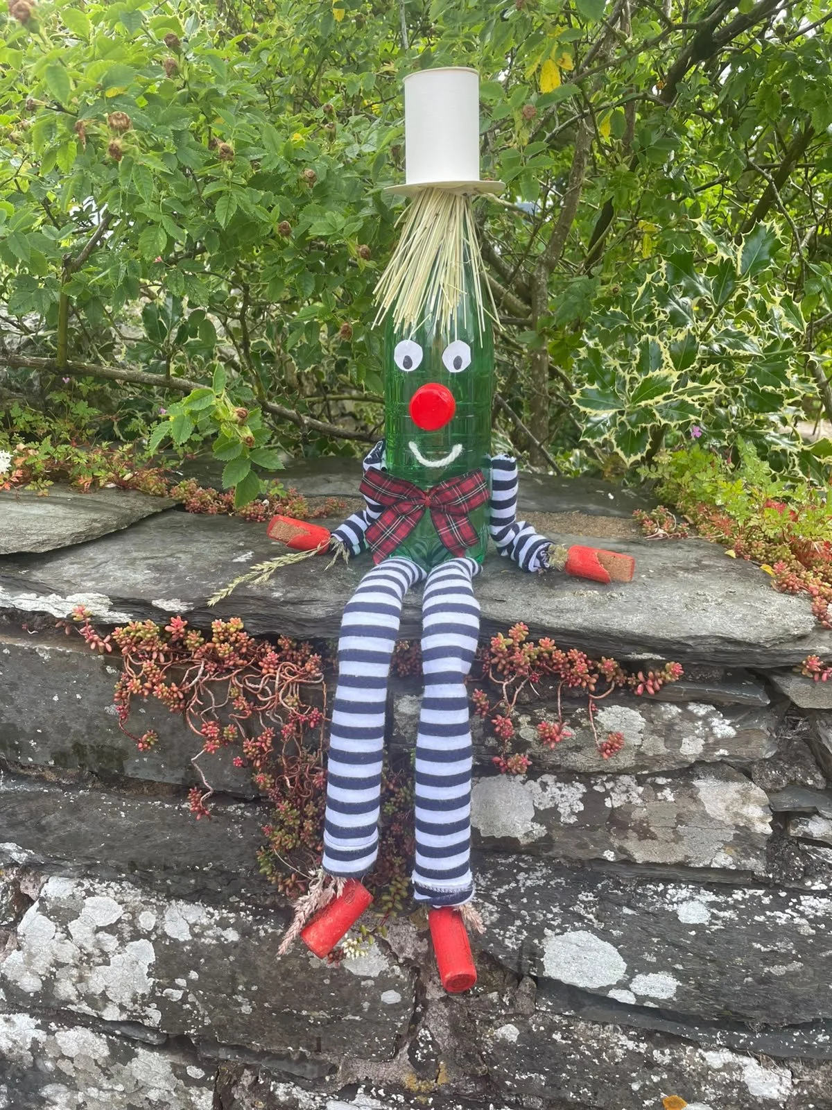 A wacky, homemade clown doll with a bottle body, striped limbs, a red nose, and a top hat, sitting on a stone wall with green bushes in the background.