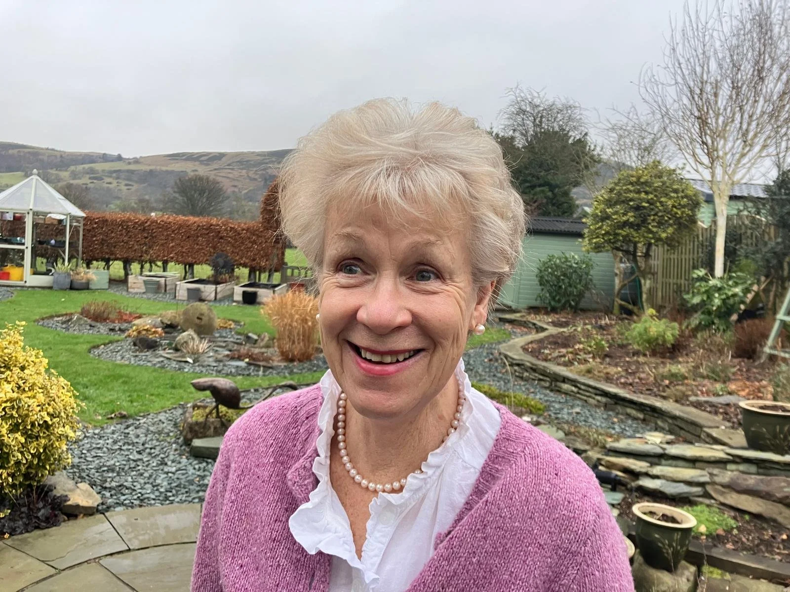 An elderly woman with short white hair smiling outdoors in a garden. She wears a pink cardigan, a white ruffled blouse, and a pearl necklace. The garden has various plants, a small pond, and a greenhouse.