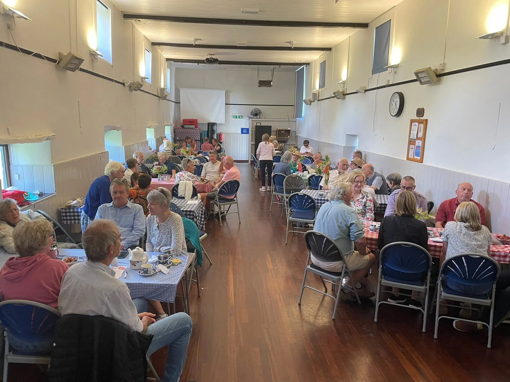 A large group of people sitting at tables with checkered tablecloths in a well-lit room, engaging in conversations and enjoying a meal.
