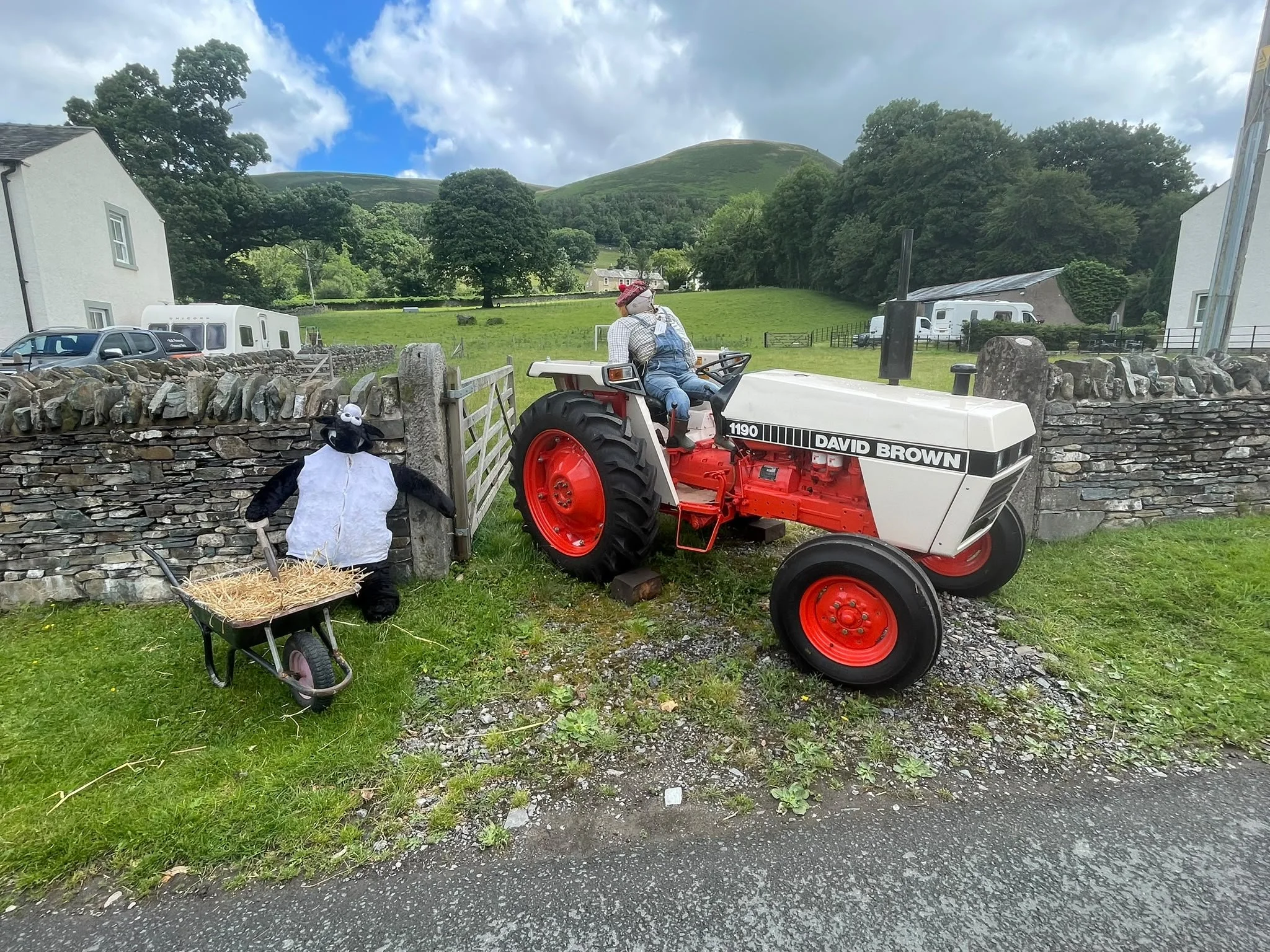 A rural landscape with green rolling hills, trees, and a cloudy sky, viewed behind a stone fence. In the foreground, there is a red and white tractor with a person sitting on it, wearing a hat. To the left, there is a black stuffed animal and a small