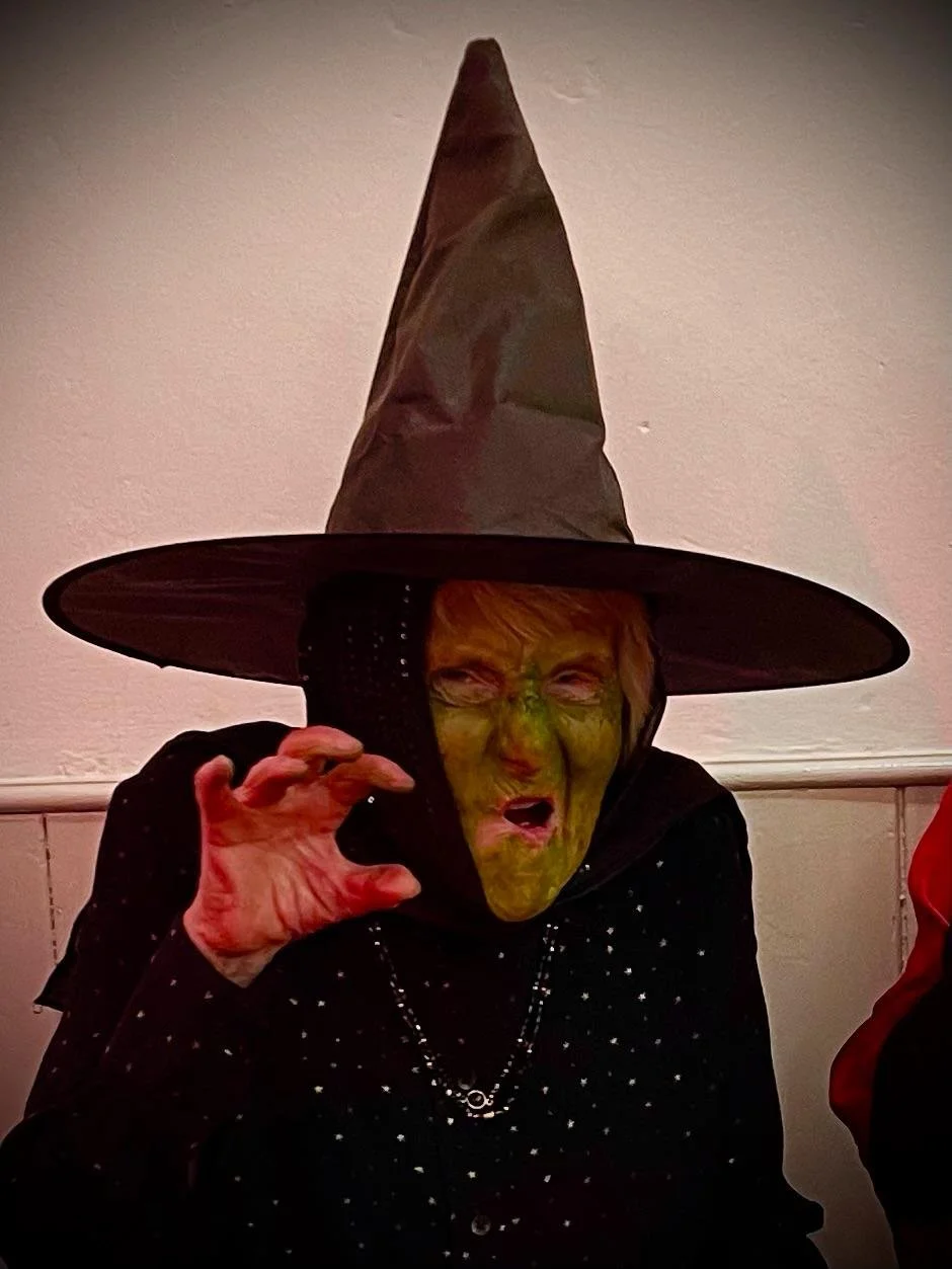 Person dressed as a witch with green face paint, wearing a large pointed black hat, black hoodie, and jewelry, making a claw gesture and showing a fierce expression.