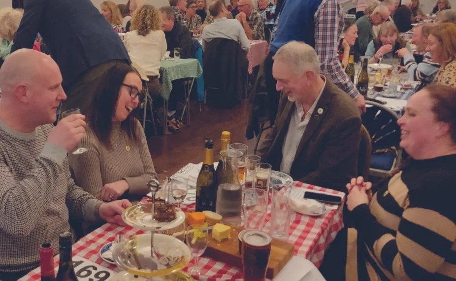 People enjoying a meal at a lively table during a gathering or celebration, with drinks, food, and a cheerful atmosphere.