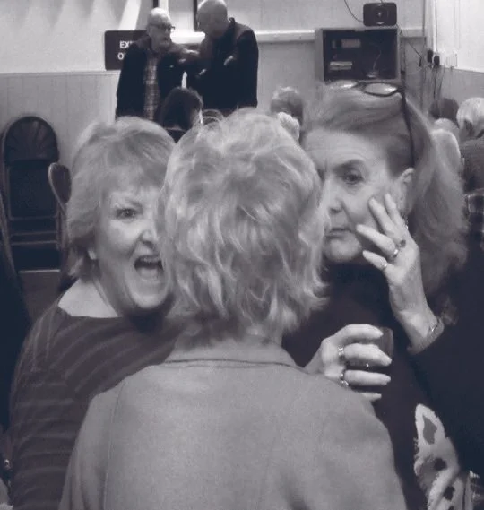 A group of women are gathered close together, engaged in conversation, with two people dancing in the background at what appears to be a social gathering or party.