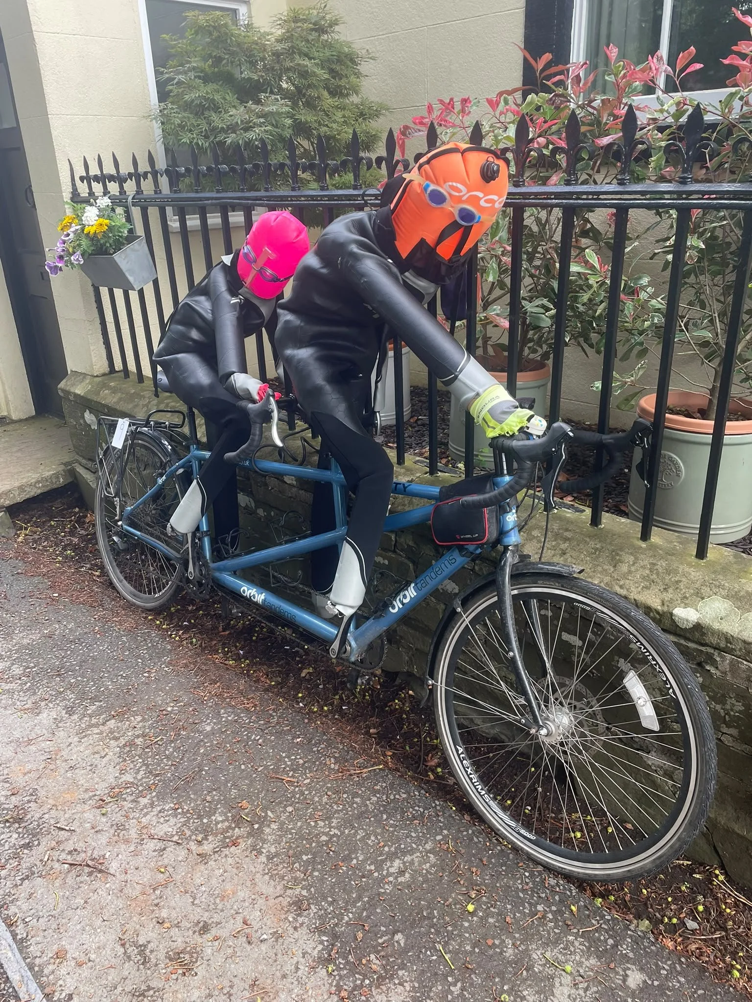 A tandem bicycle with two riders wearing wetsuits, helmets, and goggles, parked near a stone curb and a black metal fence with potted plants.