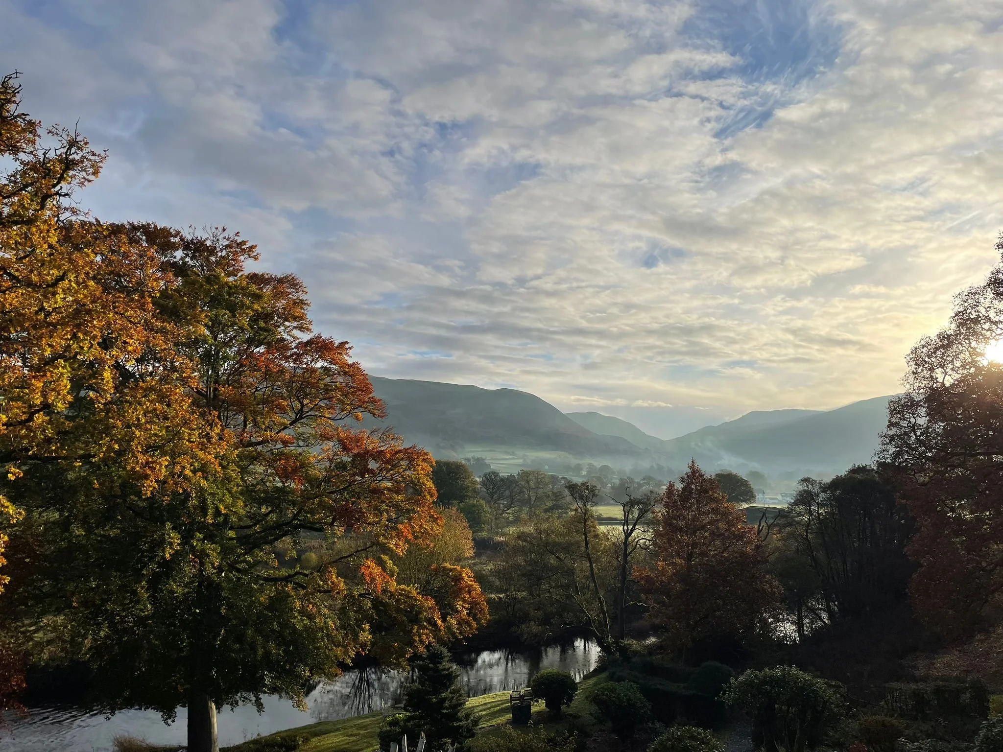 Scenic view of a lake surrounded by trees with colorful fall foliage, mountains in the distance, and a partly cloudy sky during sunrise or sunset.