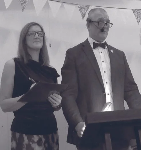 A man and a woman standing side by side at an event. The man is wearing glasses, a tuxedo with a bow tie, and a headset microphone. The woman is wearing glasses and a sleeveless dress, holding a clipboard. Bunting decorations are hanging in the backg