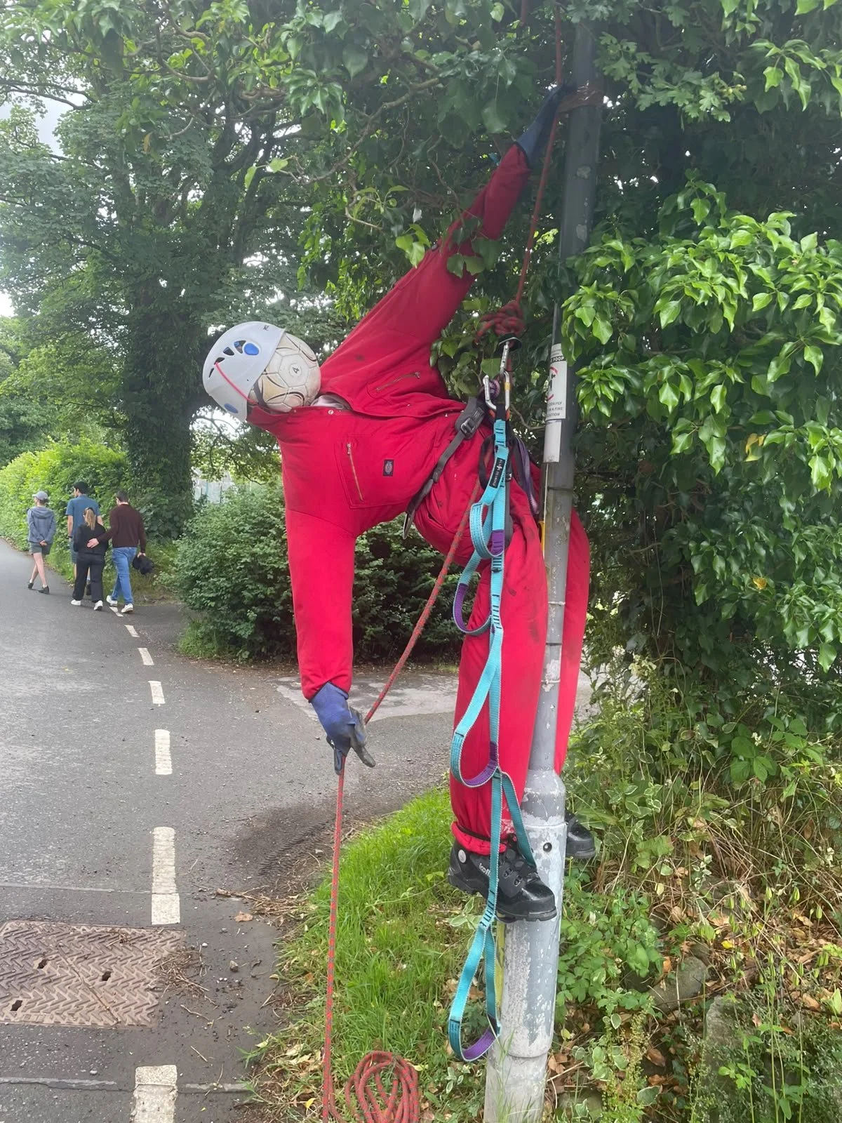 A mannequin dressed in red climbing gear, including a helmet and harness, is climbing a lamppost on a sidewalk in a park. The lamppost is surrounded by green foliage, and several pedestrians are walking nearby.
