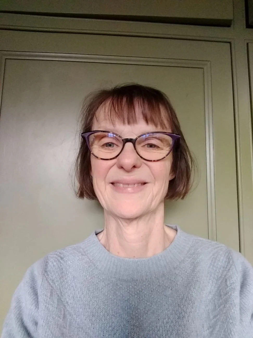 A woman with short brown hair, glasses, and a friendly smile standing in front of a green cabinet.
