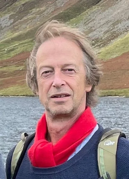 A man with gray hair and a red scarf standing outdoors near a body of water and a hillside in the background.