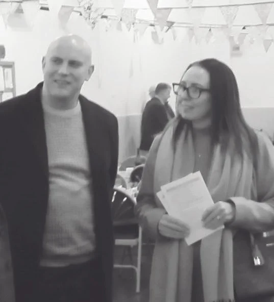 Two people, man and woman, standing indoors at an event decorated with banners, with other attendees in the background. The woman is holding papers.