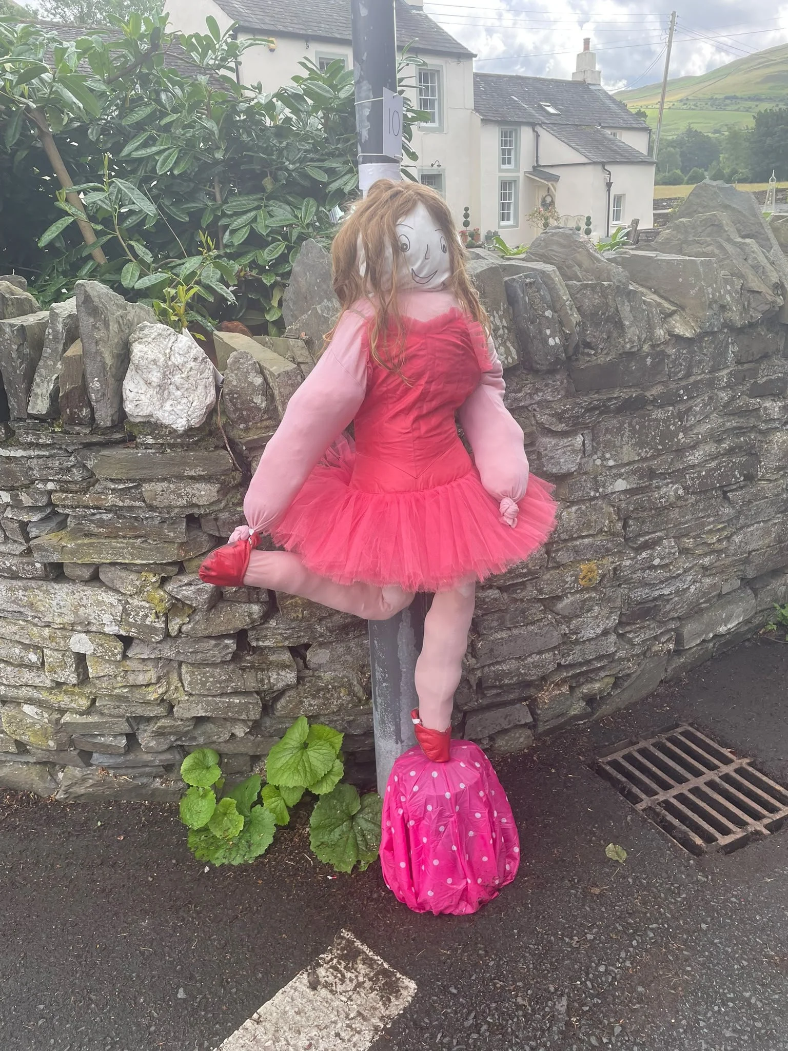 A scarecrow with a white face and drawn smile, dressed in a pink dress with puffy pink sleeves, pink socks, and pink shoes, leaning against a stone wall in a rural outdoor setting.
