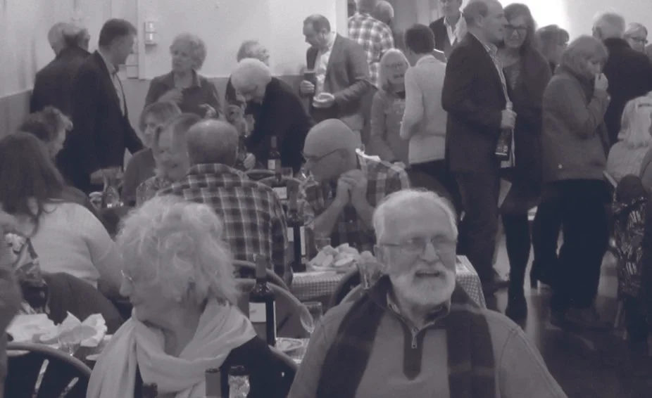 People gathered at a social event, sitting at tables and socializing, some standing and holding drinks.
