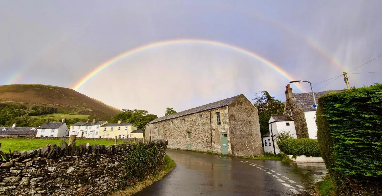 A rainy day scene in a small village with a rainbow arching over the houses and rolling hills in the background. Wet road, stone wall, and lush green hedges are visible.