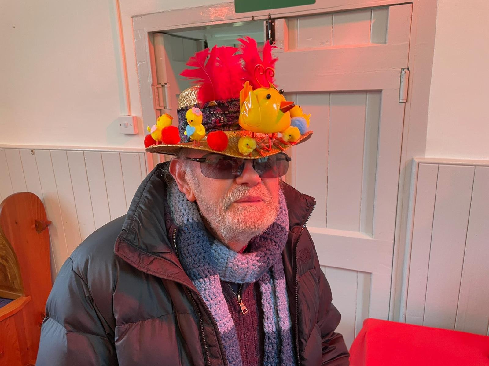 Older man wearing sunglasses, a black jacket, and a gray scarf, sitting indoors in front of a wooden door with white paneling. He is wearing a colorful hat decorated with a rubber duck, feathers, small chicks, and other decorative items.
