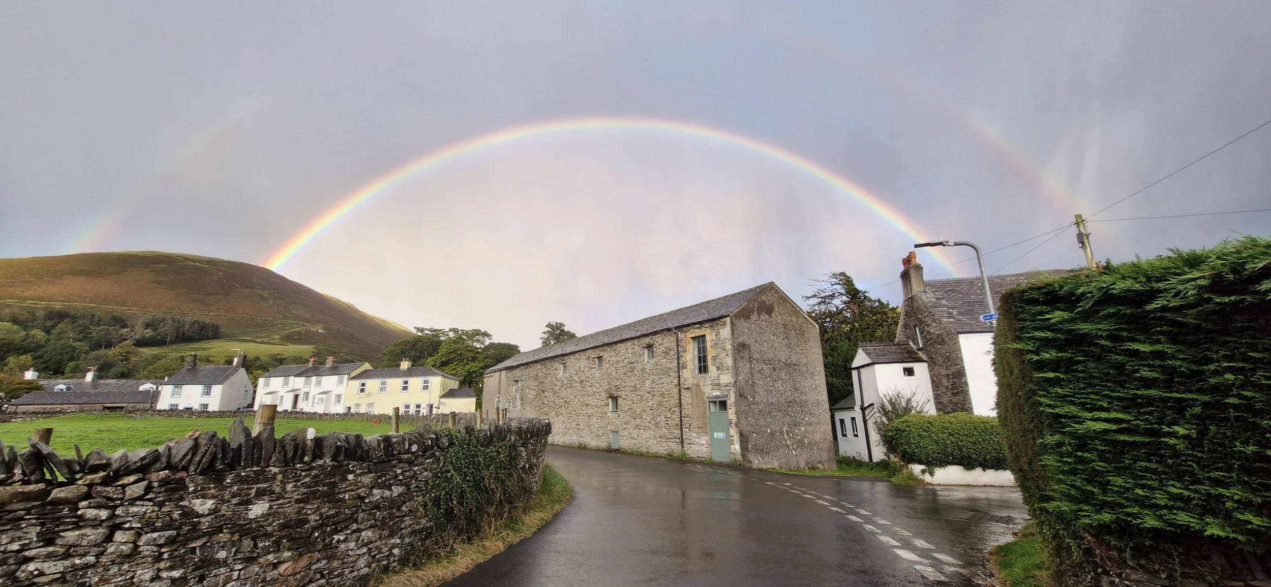 A rural street scene with a stone wall on the left, green fields, and white houses with black roofs. A stone building is on the right side of the street, and a large green hedge lines the right edge. A double rainbow arches across the cloudy sky over the landscape.