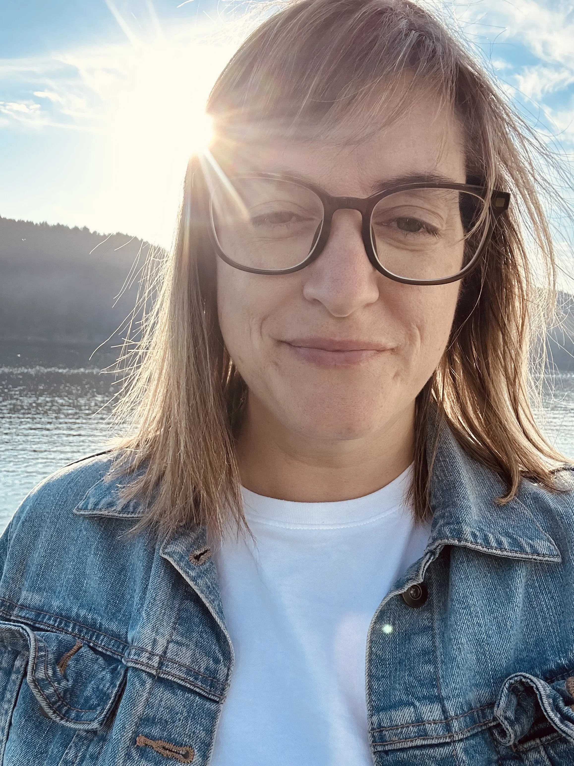 Krisi Johnson, taking a selfie outdoors near a body of water with the sun shining behind her and mountains visible in the background.