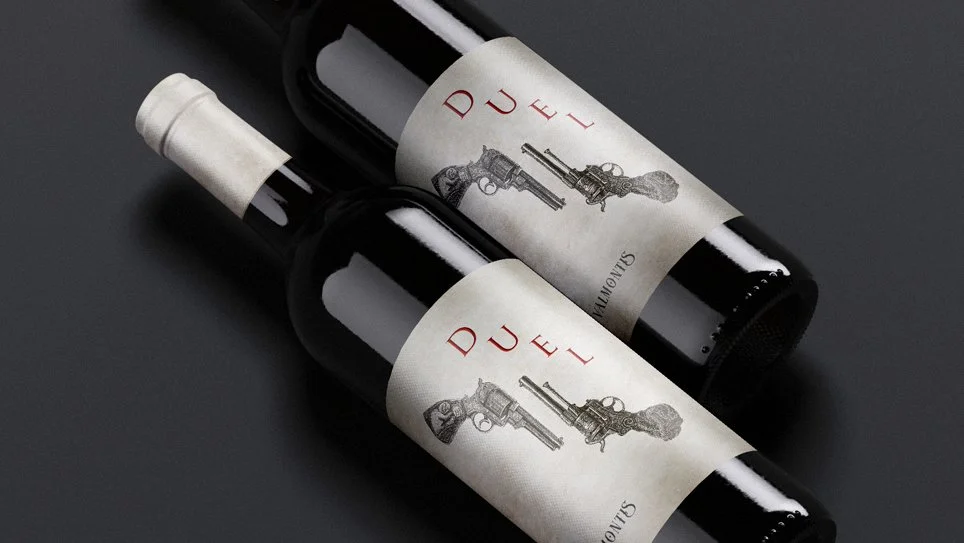 Bottle mockup “DUEL – Two Bottles” by Daniel Perez Cabrera: ink illustration of vertically inverted revolvers in symmetrical composition, expressing sobriety and tension through hand-drawn lines and classical references.