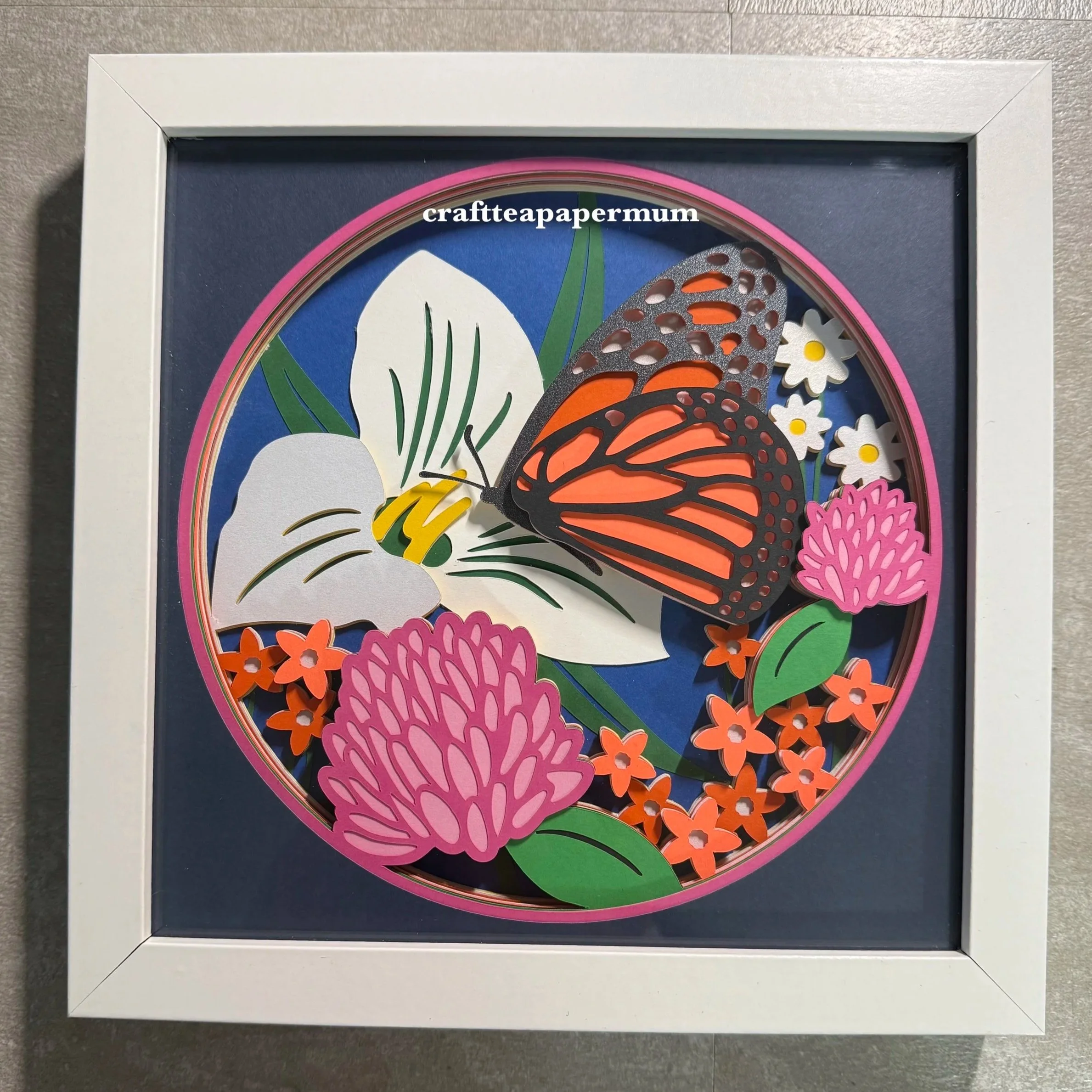 Colorful paper craft of a butterfly with pink, orange, and brown wings, surrounded by various paper flowers and leaves, framed in a white border.