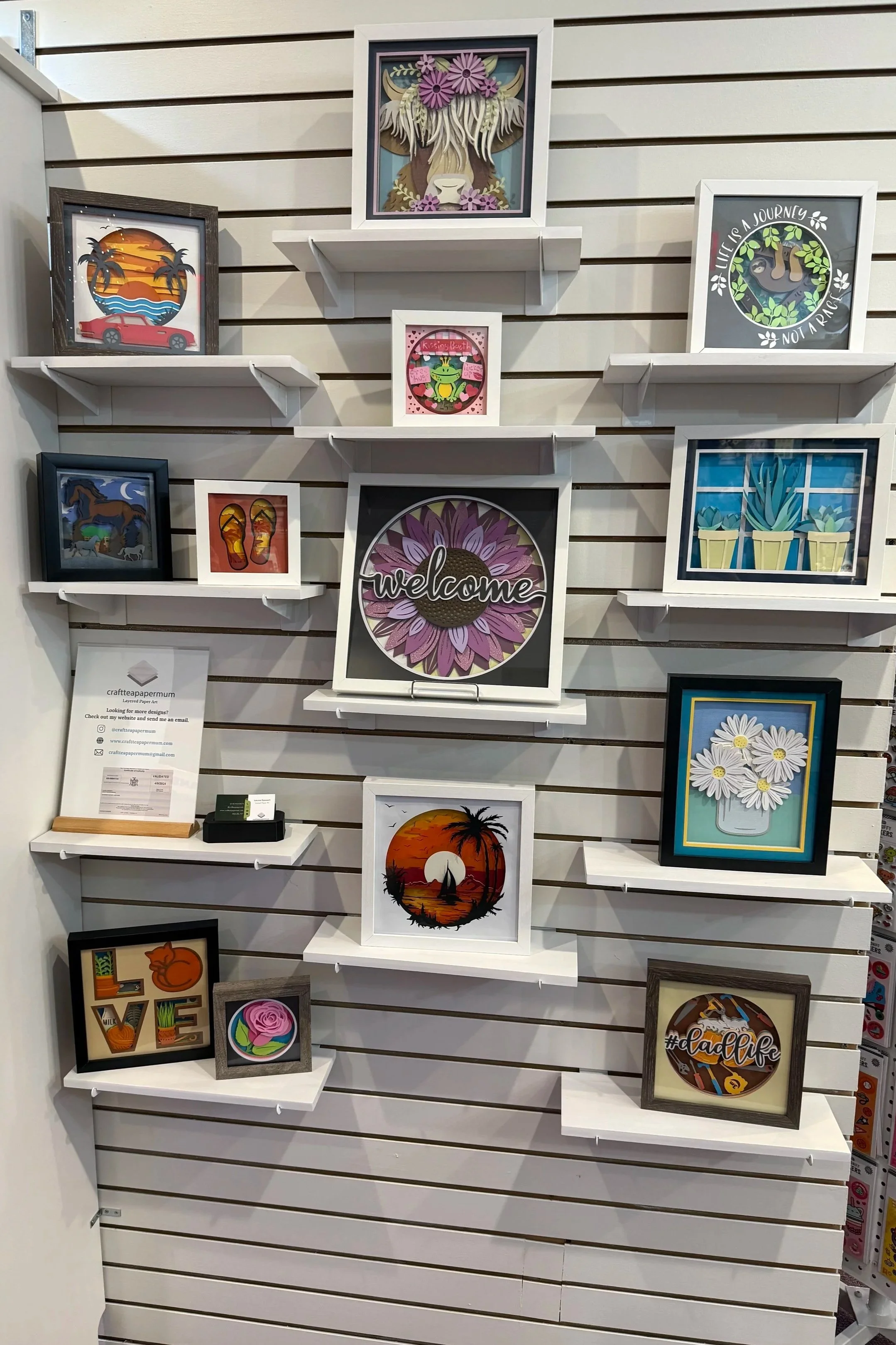 Display of framed art and decorative pictures on a slat wall in a retail setting, including scenes with sunsets, animals, plants, and abstract designs.