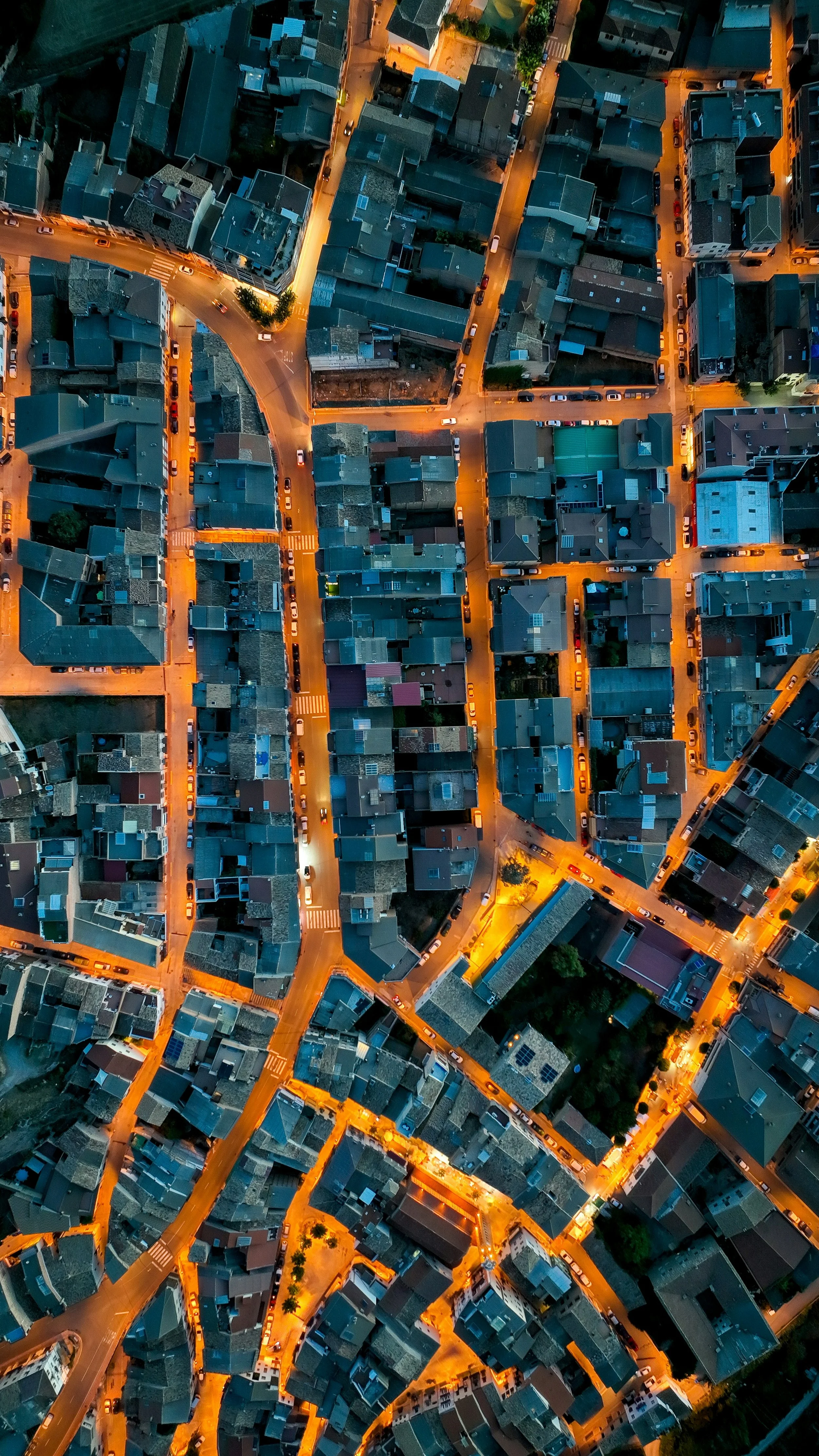 Aerial view of a city neighborhood during dusk, showing tightly packed buildings, narrow streets, and warm street lighting.