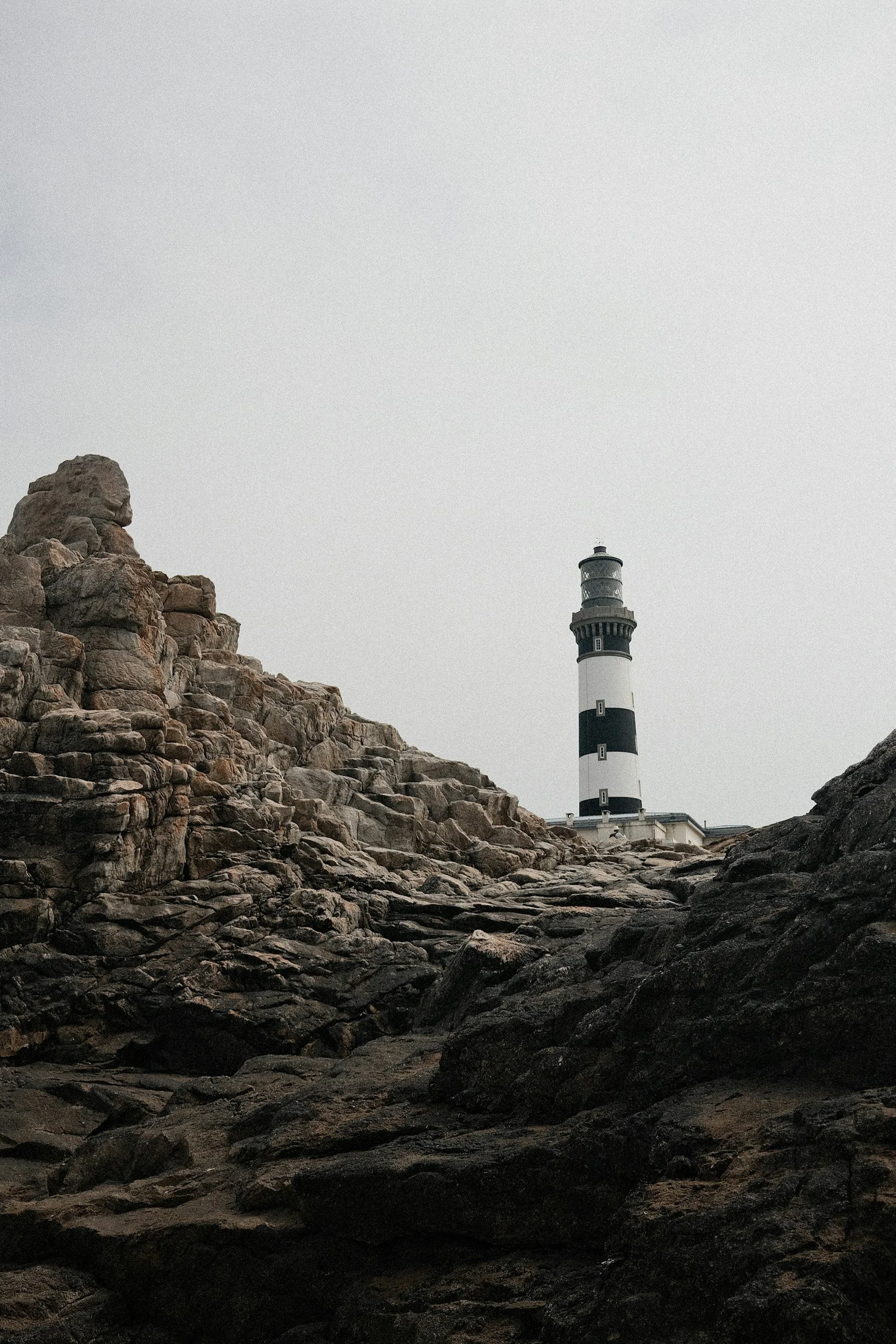 A lighthouse with black and white horizontal stripes on a rocky coastline under an overcast sky.