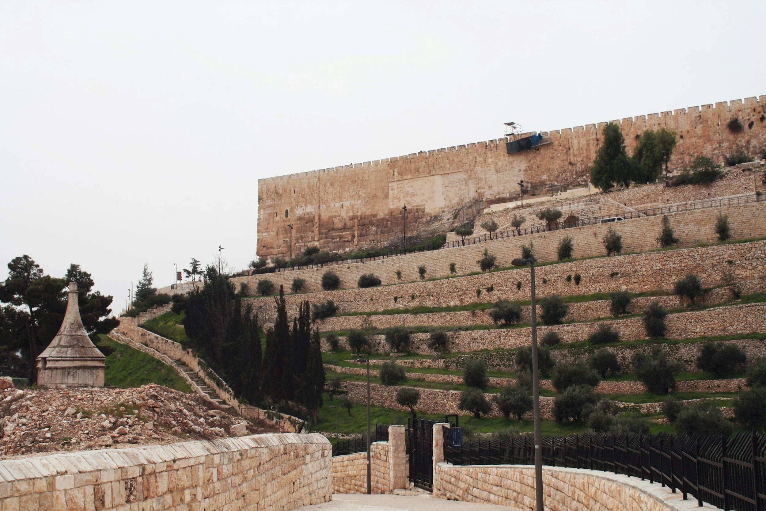 A large stone fortress with terraced landscaping filled with small trees and bushes, and a stone wall and gate at the bottom.