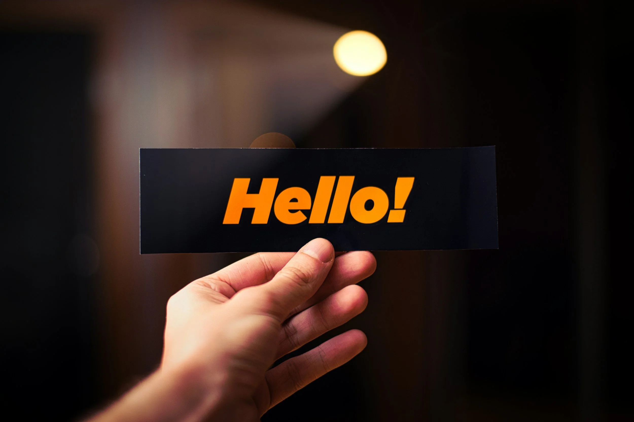 A hand holding a black sign with the word 'Hello!' in bold orange letters. The background is out of focus with warm lighting.