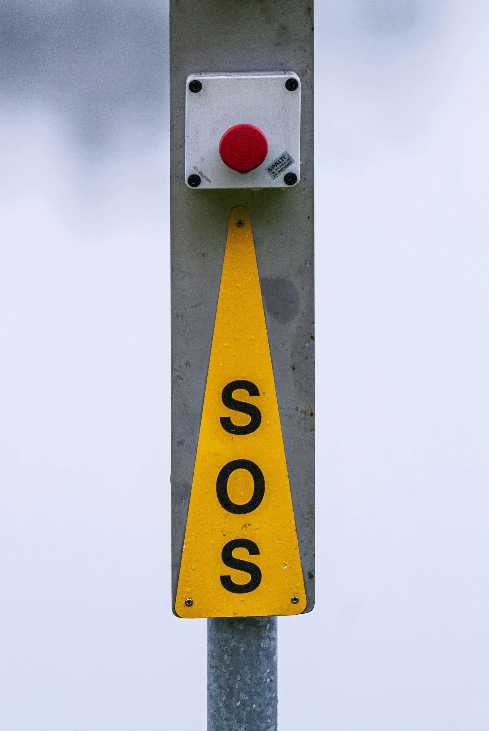 A yellow sign with black letters spelling 'S O S' hanging on a metal pole, with a red emergency button above it.