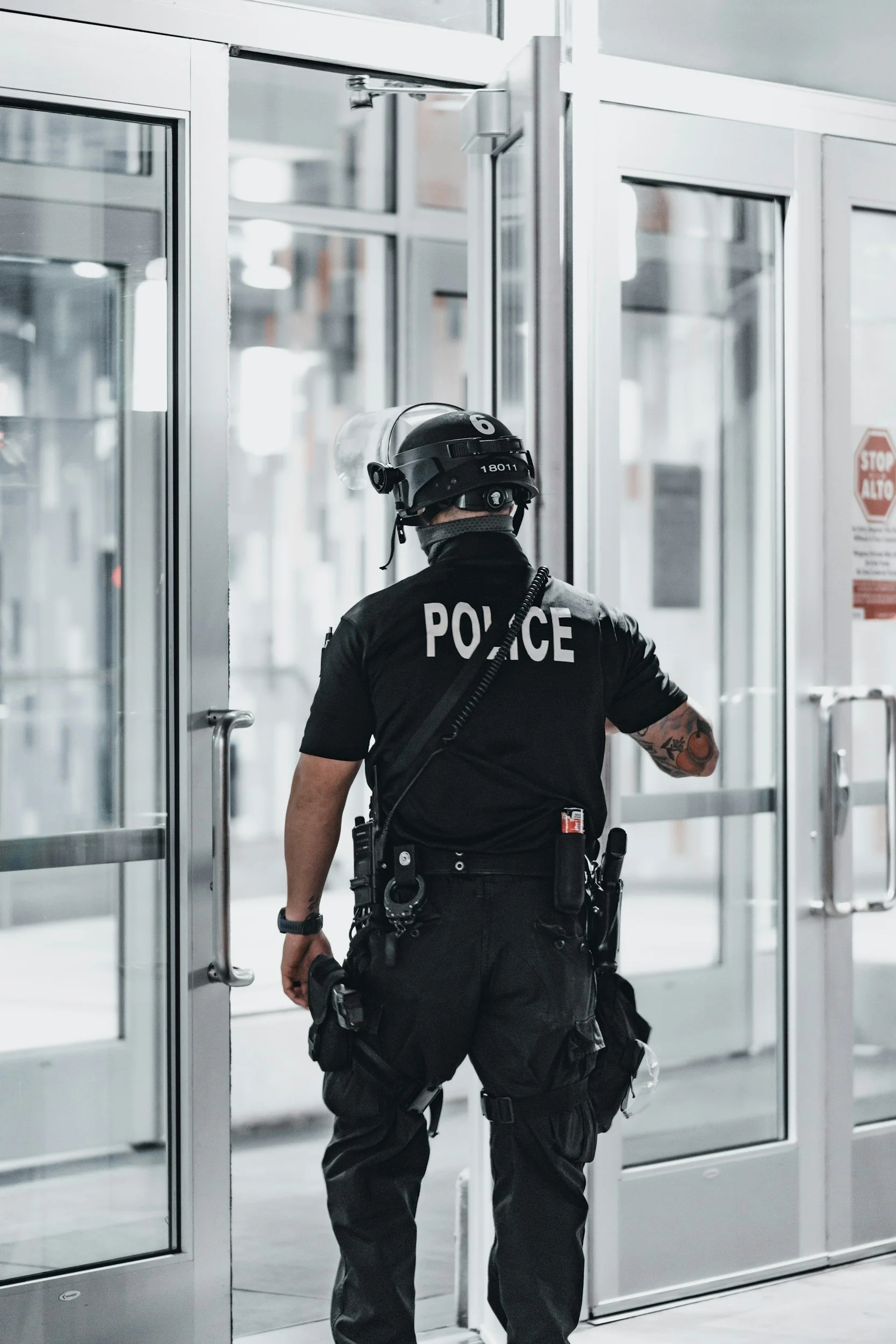 A police officer wearing a helmet and uniform, facing a glass door in an indoor setting.