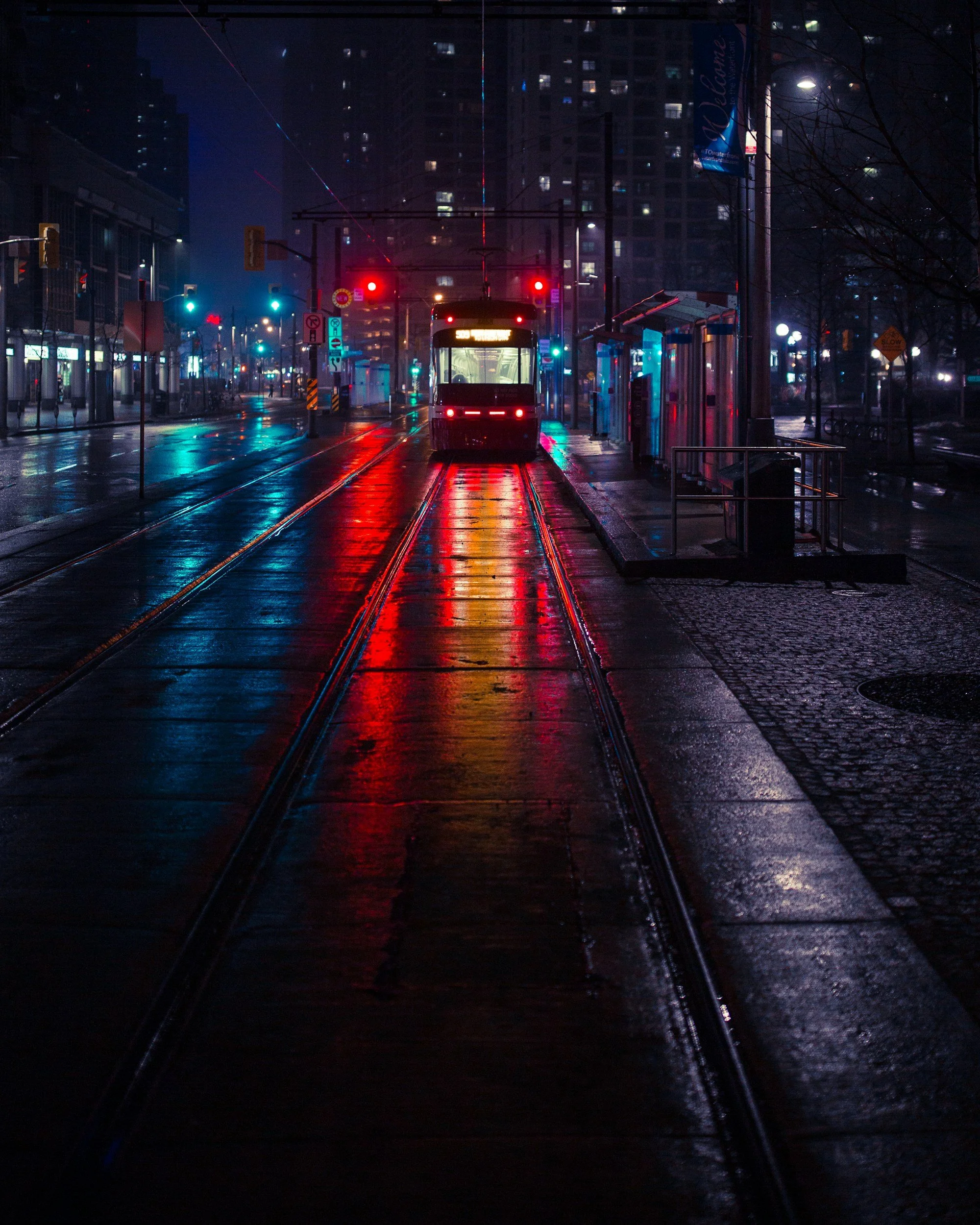 A city street at night with tram tracks, a tram stopped at a station, wet pavement reflecting colorful lights, illuminated signage, and tall buildings in the background.
