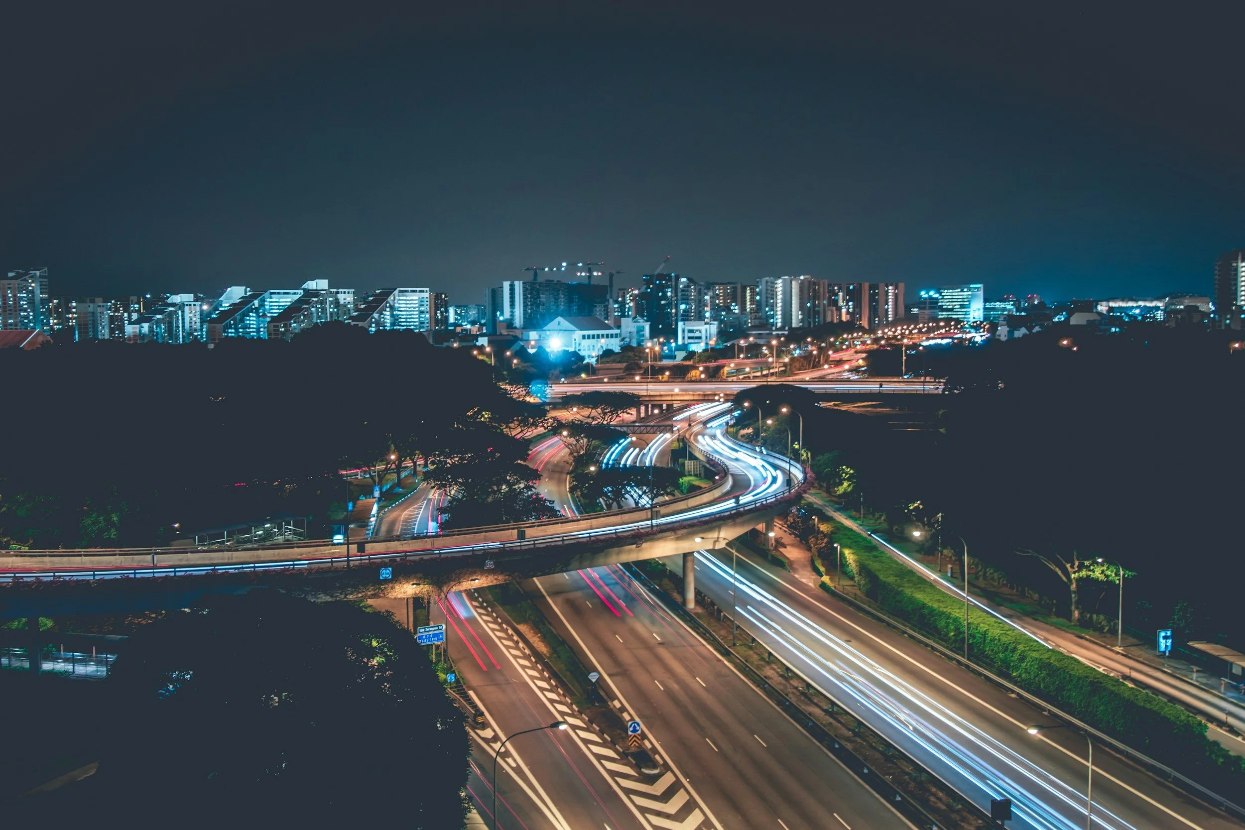 Nighttime cityscape featuring illuminated high-rise buildings, curved highway with light trails from moving vehicles, and dark sky.