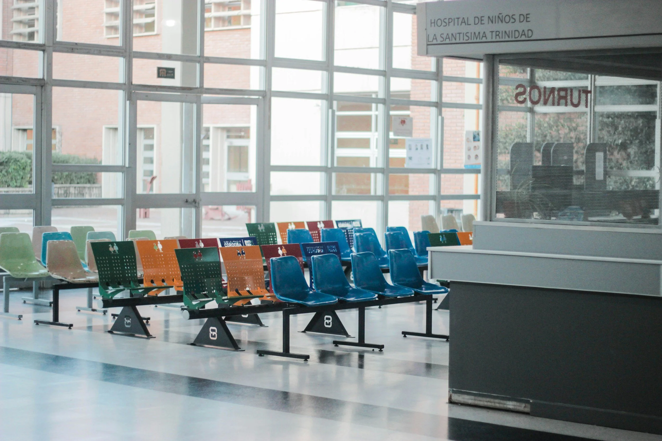 Empty hospital waiting area with multicolored chairs arranged in rows, large windows, and a reception desk on the right, with a sign indicating it is the children's hospital of La Santísima Trinidad.