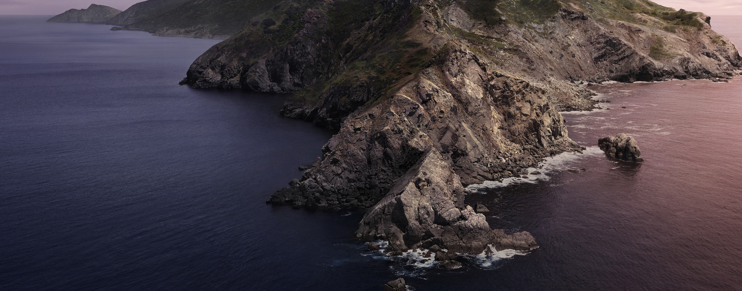 Coastal rocky cliffs extend into the ocean, with the water changing from dark blue to pinkish hues near the shore, under a pink-tinted sky.