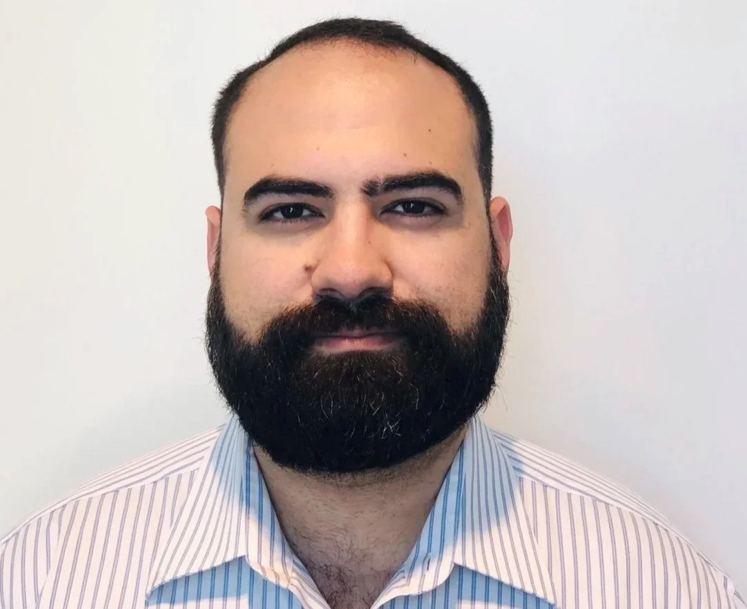 A man with a beard and short dark hair wears a collared shirt and stands against a plain white wall.