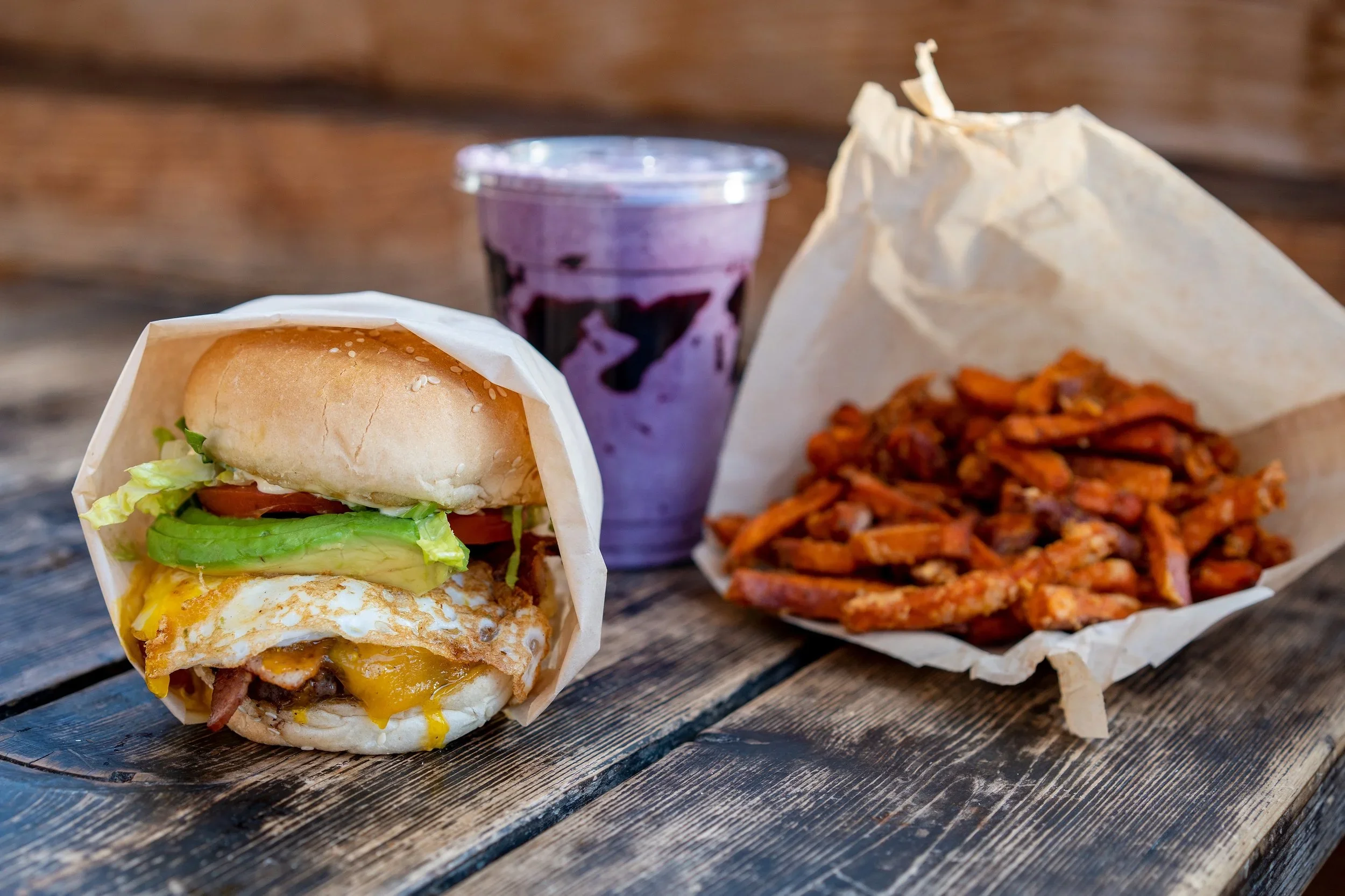 A cheeseburger with lettuce, tomato, and fried egg in a bun, a side of spaghetti with tomato sauce, and a purple berry smoothie in a plastic cup on a wooden table.