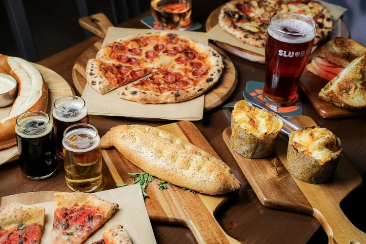 A variety of food and drinks on a dark wooden table, including pizza slices, a whole pizza, bread, stuffed baked potatoes, beer, and a glass of rosé wine.