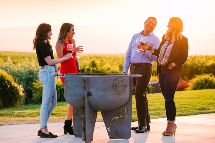 Four people holding glasses of wine standing outdoors near a large outdoor grill during sunset.